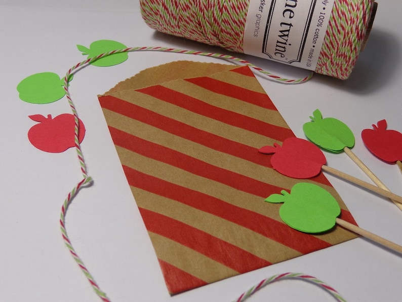 May include: A brown paper bag with red and white stripes. The bag is decorated with green and red paper apple cutouts. The bag is sitting on a white surface with a spool of red and white twine.