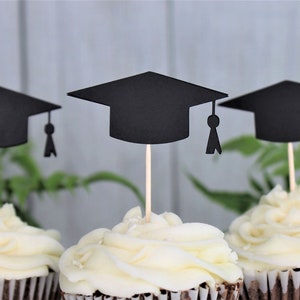 May include: Three cupcakes with black graduation cap toppers. The cupcakes are frosted with white frosting and have brown cupcake liners.