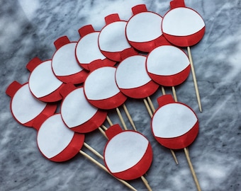 Puede incluir: Un conjunto de 12 adornos para cupcakes de flotador de pesca rojo y blanco. Los flotadores están hechos de papel y tienen la parte superior blanca y la inferior roja. Están unidos a brochetas de madera.