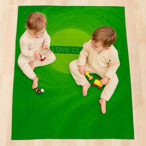 Puede incluir: Dos bebés con monos beige sentados sobre una alfombra de juego verde con el texto "LOVE CL". La alfombra tiene un diseño circular en tonos verdes. Cada bebé sostiene un juguete, uno rojo y negro, el otro amarillo y rojo.