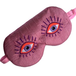 May include: A pink sleep mask with embroidered eye designs. The eyes have orange irises, purple pupils, and purple eyelashes. The mask has a pink strap for securing it around the head.
