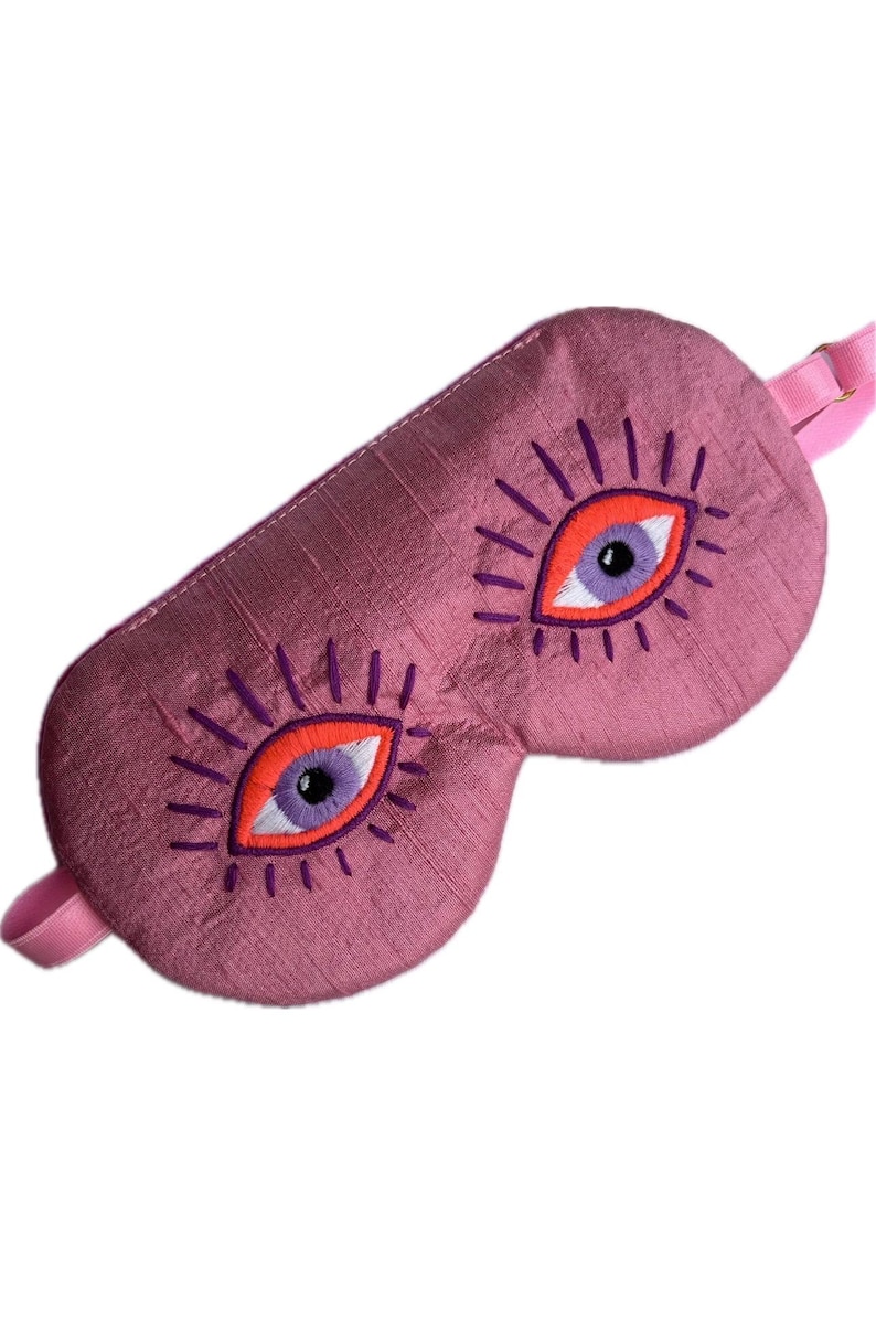 May include: A pink sleep mask with embroidered eye designs. The eyes have orange irises, purple pupils, and purple eyelashes. The mask has a pink strap for securing it around the head.