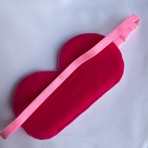 May include: A heart-shaped sleep mask in a vibrant fuchsia color, with a matching pink elastic band. The mask is made of a soft, velvety material, designed for comfort and light blockage. The elastic band is attached with small gold-colored hardware.