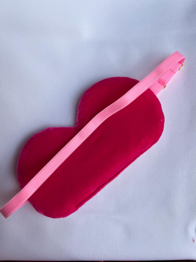May include: A heart-shaped sleep mask in a vibrant fuchsia color, with a matching pink elastic band. The mask is made of a soft, velvety material, designed for comfort and light blockage. The elastic band is attached with small gold-colored hardware.