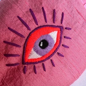 May include: Pink sleep mask with an embroidered eye design. The eye features a red outline, a purple iris, a white highlight, and black pupil. Purple lines radiate from the eye, and the mask has a pink strap.