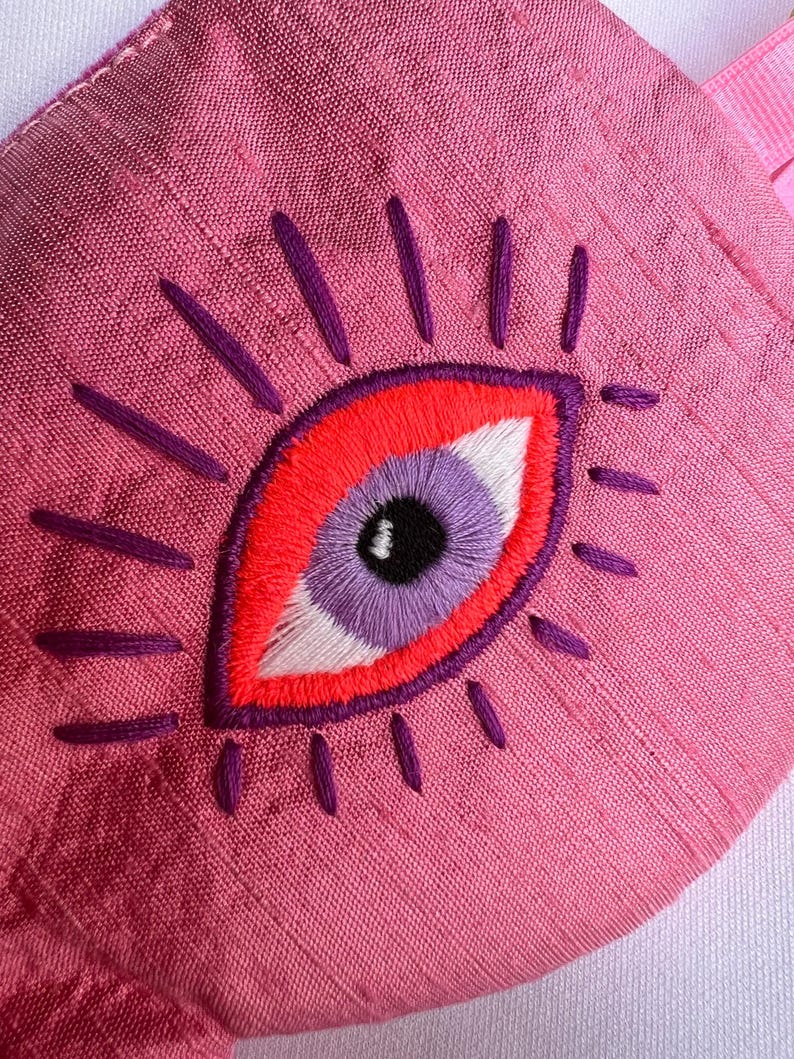 May include: Pink sleep mask with an embroidered eye design. The eye features a red outline, a purple iris, a white highlight, and black pupil. Purple lines radiate from the eye, and the mask has a pink strap.