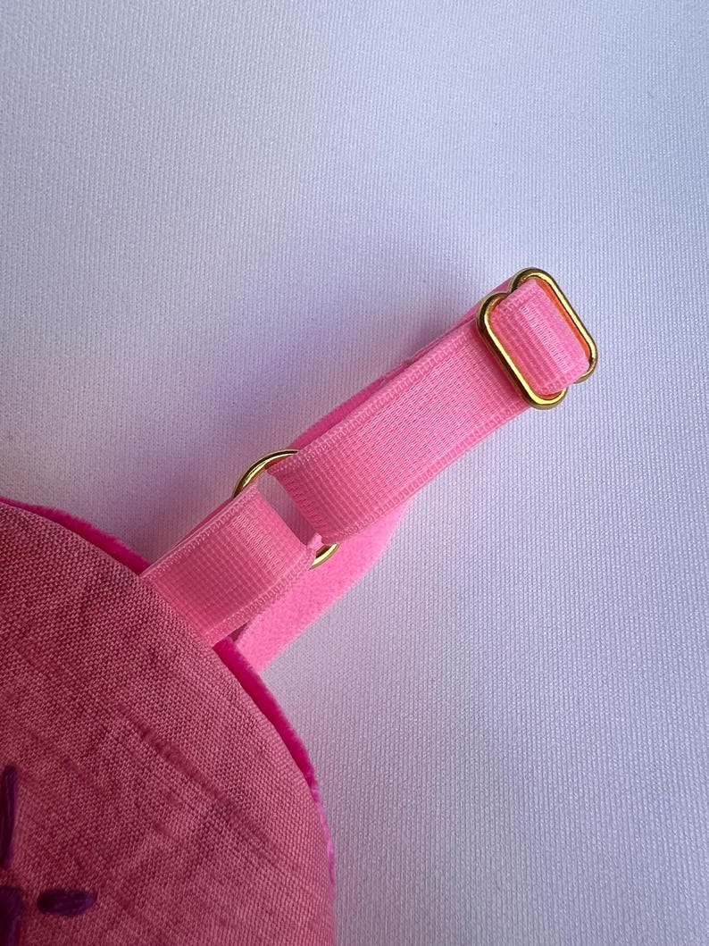 May include: Close-up of a pink fabric item with a matching pink strap and gold-colored metal hardware. The strap is attached to the item with a gold ring. The item appears to be made of a textured fabric.
