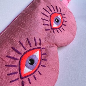 May include: Pink sleep mask with embroidered eye design. The mask features two large eyes with red, purple, and white details, surrounded by purple radiating lines. It has a pink adjustable strap with a gold-colored buckle.