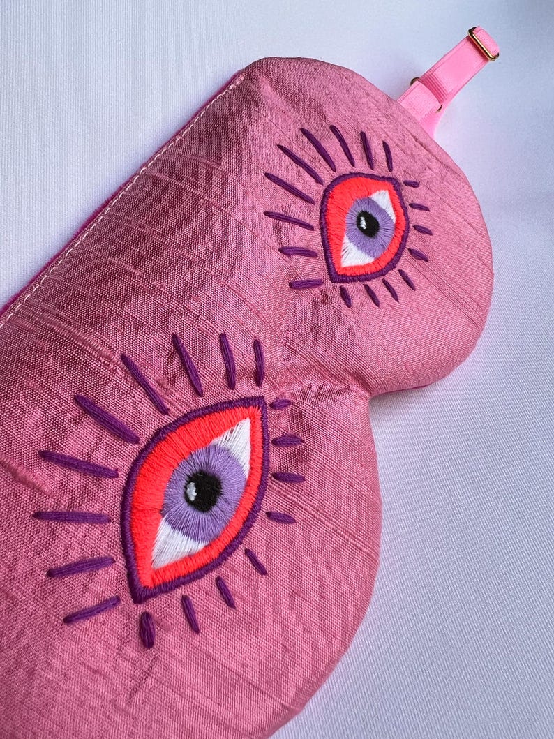 May include: Pink sleep mask with embroidered eye design. The mask features two large eyes with red, purple, and white details, surrounded by purple radiating lines. It has a pink adjustable strap with a gold-colored buckle.