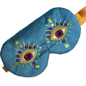 Op de afbeelding: Teal oogmasker met geborduurde gele en paarse oogdetails, geaccentueerd met groene en iriserende pailletten. Het slaapmasker heeft een gele elastische band. Een uniek en decoratief slaapaccessoire.