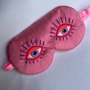 May include: Pink sleep mask with embroidered eye design. The mask features two large eyes with orange and purple details, surrounded by purple eyelashes. A pink strap is attached for securing the mask.