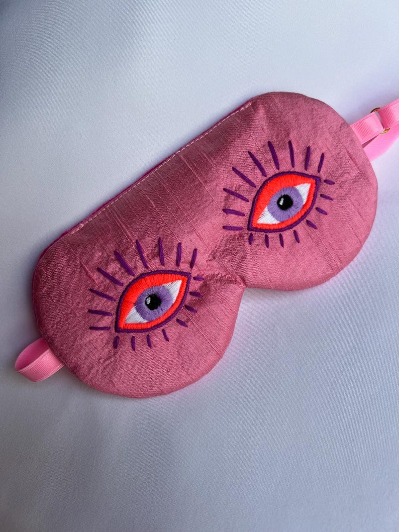 May include: Pink sleep mask with embroidered eye design. The mask features two large eyes with orange and purple details, surrounded by purple eyelashes. A pink strap is attached for securing the mask.