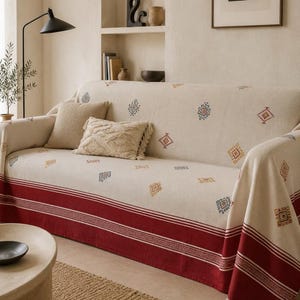 May include: A cream-coloured sofa throw blanket featuring small, scattered geometric patterns in blue, orange, and red. The blanket has a wide, solid dark red border along the bottom edge with intricate white linear detailing and a fringed hem.