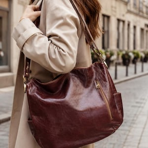 May include: A dark brown leather hobo bag with a glossy finish, featuring a vertical zippered side pocket with gold-toned hardware, a top zipper closure, and a shoulder strap attached with gold-toned metal rings, worn over a beige trench coat.