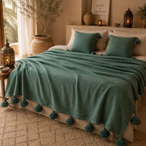 May include: A teal green textured throw blanket with matching tassels and two square cushions on a bed. The bed is covered with a cream-coloured bedspread. Decorative lanterns and a potted plant are visible in the background.