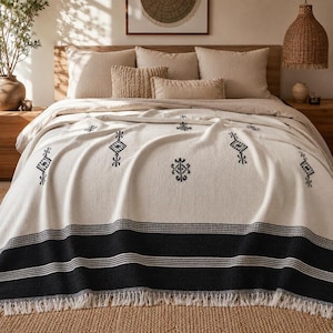 Op de afbeelding: Een crèmekleurige deken met een zwart geometrisch patroon en franjes is over een bed gedrapeerd. De deken heeft een dikke zwarte streep aan de onderkant met horizontale witte strepen. Decoratieve kussens en een houten hoofdbord zijn op de achtergrond.