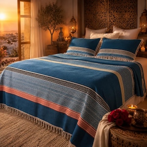 May include: A blue bedspread with a geometric pattern and fringe trim. The bedspread has stripes of white, black, and red. Two matching blue pillowcases with fringe are on the bed. The room has a warm, inviting atmosphere.