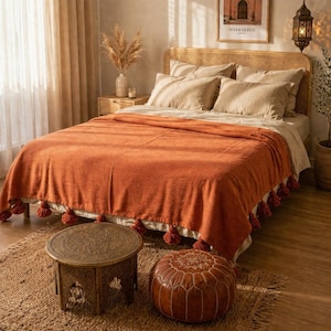 May include: A bedroom scene with a bed featuring a woven headboard and beige bedding. An orange throw blanket with tassel trim is draped over the bed. A small, round wooden table and a leather ottoman sit on a jute rug in front of the bed.