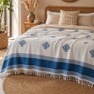 Op de afbeelding: Een wit en blauw patroon plaid met franjes. De deken heeft een blauwe rand en geometrische ontwerpen. De plaid is gedrapeerd over een bed met kussens in neutrale tinten en een geweven hoofdbord. Een ronde jute vloerkleed ligt op de grond.