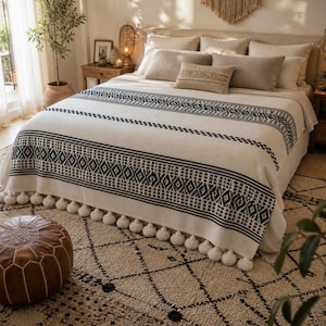May include: A cream-coloured bedspread with a black geometric pattern and a row of white pom-poms along the edge. The bed has several pillows and a decorative cushion. The bed is on a patterned rug.