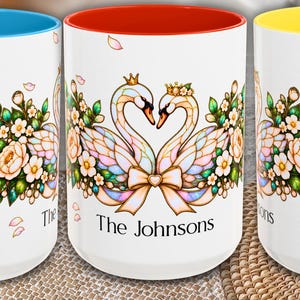 May include: Three white ceramic mugs with colored interiors and handles in blue, red, and yellow. The mugs feature a stained-glass style illustration of two swans forming a heart, floral accents, and the names Noah and Hannah, plus The Johnsons.