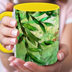 May include: A ceramic mug with a bright yellow handle and interior rim. The exterior features a green stained glass pattern of leaves and branches with the partial red text "LIFE" and "IN THE" printed on the side of the mug surface.