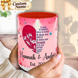 May include: A pink ceramic mug with a red interior, featuring a large red heart graphic containing white floral illustrations and the text: Love is patient love is kind It does not envy It does not boast it is not proud 1 Corin thians 13:4. Below the heart, the names Hannah & Andrew and Est. 2020 are printed.