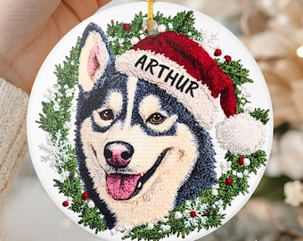May include: A round white ceramic ornament featuring a detailed illustration of a black and white husky dog wearing a red Santa hat with the name ARTHUR printed on the brim, surrounded by a green pine wreath with red berries and white snowflakes.