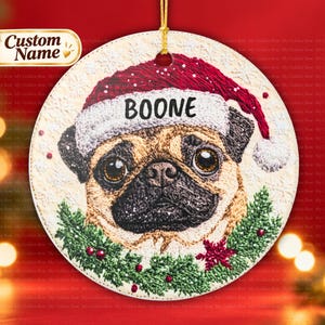 May include: A round ceramic Christmas ornament featuring a pug wearing a red and white Santa hat with the name BOONE printed in black. The pug is framed by a green pine wreath with red berries, all set against a textured cream background.