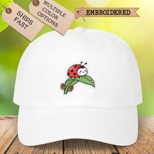 May include: A white baseball cap featuring an embroidered design of a red ladybug with black spots resting on a green leaf with small red buds. The cap has a structured crown and a curved brim, showcasing a clean and simple aesthetic.