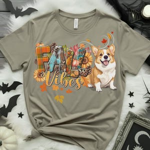 Può includere: T-shirt grigio chiaro con la scritta "FALL Vibes" in un design colorato a tema autunnale. Il design include zucche, girasoli e un cane corgi. Le lettere sono decorate con motivi a quadri, floreali e leopardati.
