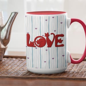 May include: White ceramic mug with a red interior and handle. The mug features the word "LOVE" in red, with cat silhouettes replacing the letters. The mug has a striped pattern with small purple hearts.