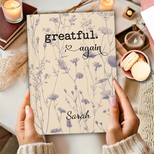 May include: A beige journal with a floral design and the words "greatful. again" and the name "Sarah" in black script. The journal is held by a person. The background includes candles, books, and pastries.