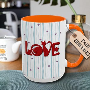 May include: White ceramic mug with an orange interior and handle. The mug features the word "LOVE" in red with cat silhouettes, and a pattern of blue vertical lines and purple hearts. A tag reads "FAST SHIPPING".