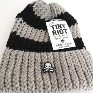 Puede incluir: Un gorro de punto a rayas grises, negras y blancas con un parche de calavera y huesos cruzados. El gorro tiene un borde acanalado y una etiqueta que dice "TINY RIOT punk rock baby gear REBEL FROM DAY ONE."