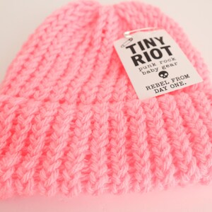 Puede incluir: Un gorro tejido de color rosa brillante con un diseño acanalado. El gorro tiene un borde doblado y una etiqueta que dice "TINY RIOT punk rock baby gear REBEL FROM DAY ONE."
