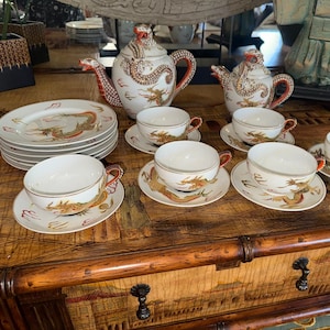 Puede incluir: Un juego de té completo con tazas, platillos, platos y teteras de porcelana blanca. El juego está decorado con un motivo de dragón en oro, rojo y marrón. Las teteras tienen picos y asas con forma de dragón. El juego se exhibe sobre una superficie de madera.
