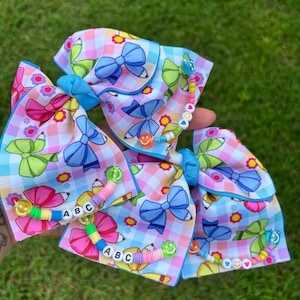May include: A hair bow made of light blue and white checkered ribbon featuring a pattern of colourful bows, flowers, and pencils. The bow is embellished with strands of multi-coloured beads, including letter beads spelling ABC and small smiley face beads.