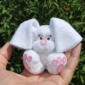 Puede incluir: Un conejito de toalla blanco con orejas, nariz y huellas de patas rosas. El conejito tiene ojos bordados y una expresión dulce.