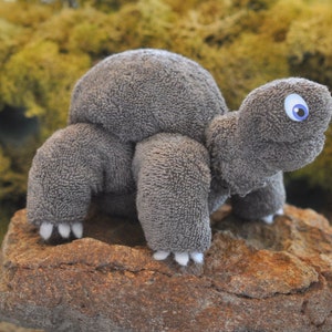 May include: A gray plush toy turtle with a blue eye, sitting on a brown rock. The turtle is made of a soft, fuzzy material.
