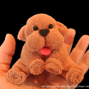 Puede incluir: Un juguete de peluche de perro marrón con ojos negros y una lengua rosa. El perro está sentado en la mano de una persona.