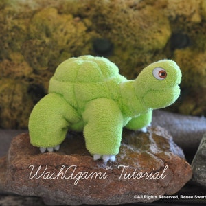 May include: A green washcloth turtle toy with white claws and a red eye. The toy is sitting on a brown rock. The text "Wash'agami Tutorial" is written on the rock.