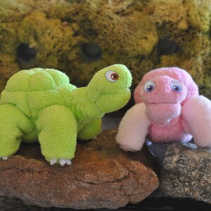 May include: Two handmade plush toys shaped like turtles. One is green and the other is pink. Both have googly eyes.
