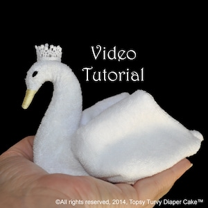 May include: A white towel folded into the shape of a swan with a small white crown on its head. The swan is being held in a person's hand. The text "Video Tutorial" is written in white on a black background.