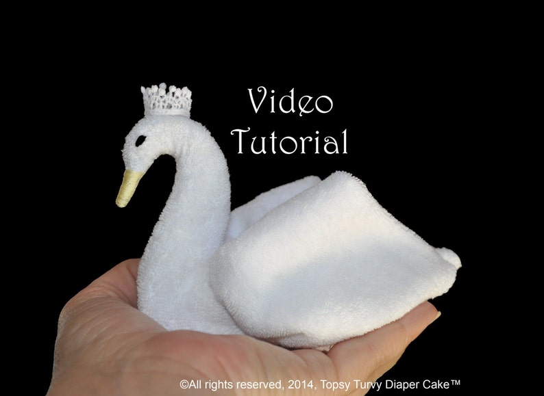 May include: A white towel folded into the shape of a swan with a small white crown on its head. The swan is being held in a person's hand. The text "Video Tutorial" is written in white on a black background.