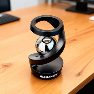 May include: A black desk ornament with a spiral design, holding a reflective silver sphere. The base of the ornament has the name "ALEXANDER" printed in white. The ornament is on a wooden desk.
