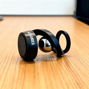 May include: A black magnetic phone holder with the name "ALEXANDER" printed on the side. The holder features a curved design with a silver metal ball in the center. It is resting on a wooden surface.