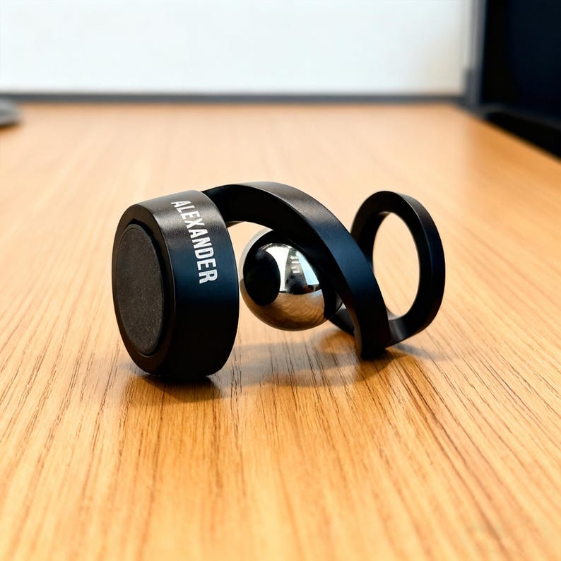 May include: A black magnetic phone holder with the name "ALEXANDER" printed on the side. The holder features a curved design with a silver metal ball in the center. It is resting on a wooden surface.