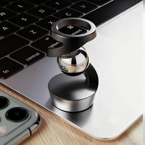 Op de afbeelding: Een zwart en zilveren telefoonstandaard met een ringgreep, een bolvormige bal en een cirkelvormige basis. De standaard staat op een laptoptoetsenbord en een smartphone is zichtbaar op de achtergrond.