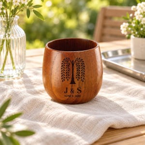 May include: A rounded wooden cup with a smooth, polished finish featuring a dark brown engraved willow tree graphic above the text J & S and the words SINCE 2011. The cup has a wide, open top and a curved body with visible wood grain patterns.